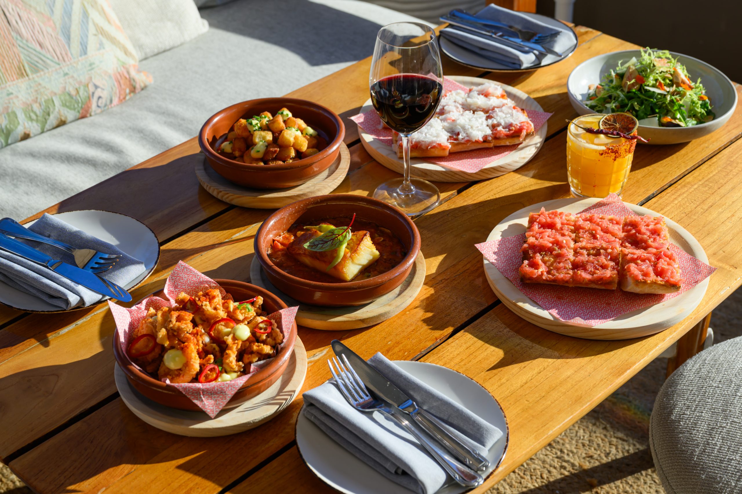 Assortment of Spanish tapas, red wine, and a cocktail served on a sunlit wooden table.